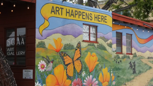 A wall painting on a side of a building features yellow with black lettering that says, "Art Happens Here." In the lower half, the painting shows an orange butterfly surrounded by orange and pink colored flowers.